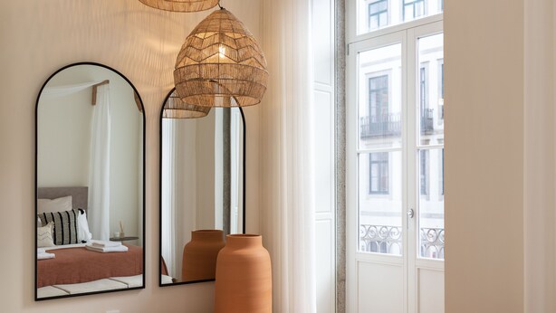 Habitación de hotel con cama, dos espejos, ventana con vista a edificios y dos jarrones grandes sobre la mesita de noche.