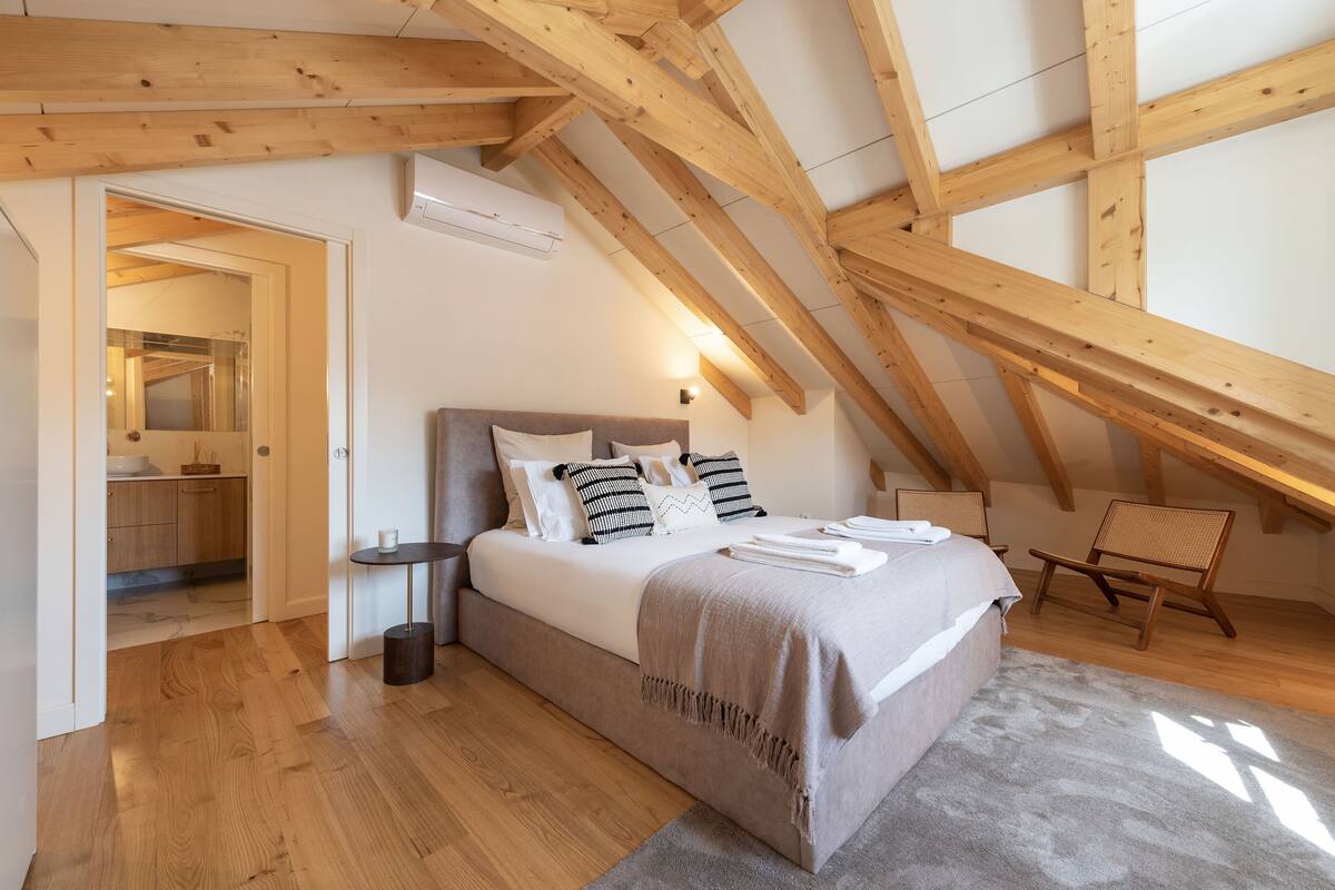 A bedroom with a wooden ceiling, a bed with a grey blanket, a bedside table, and a chair.