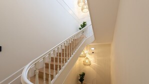 Una escalera de caracol con barandillas blancas y peldaños de madera, que conduce a un rellano con una planta en maceta y una lámpara fijada a la pared.