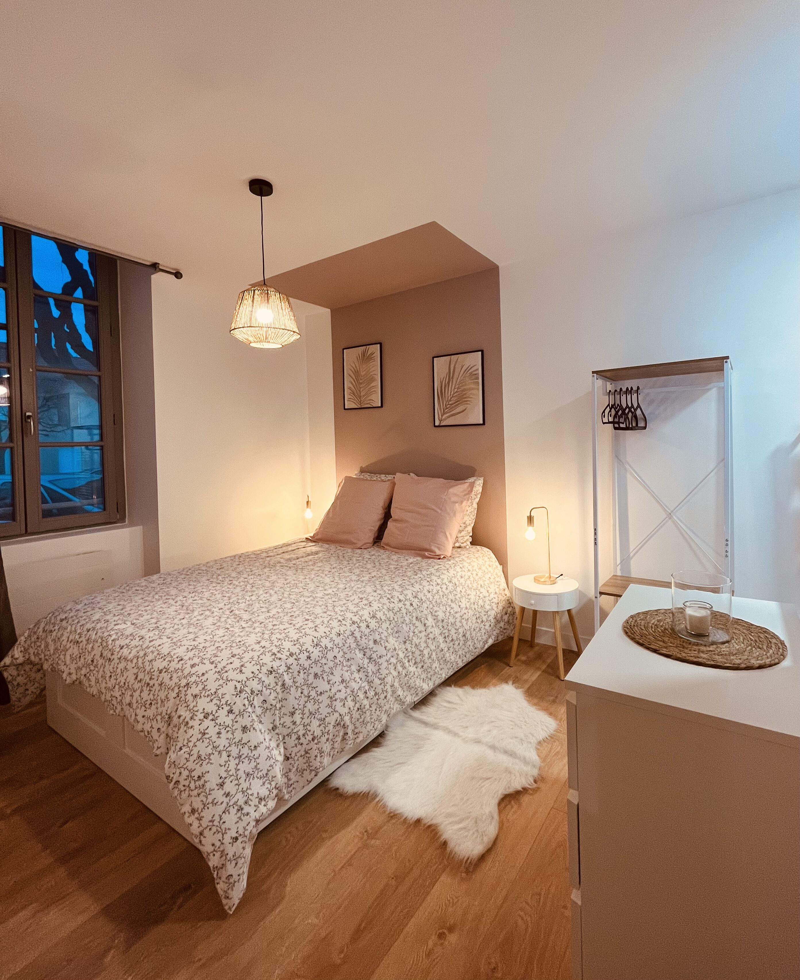 A bedroom with a bed, a nightstand, a lamp, a window with a view, and a hanging coat rack.