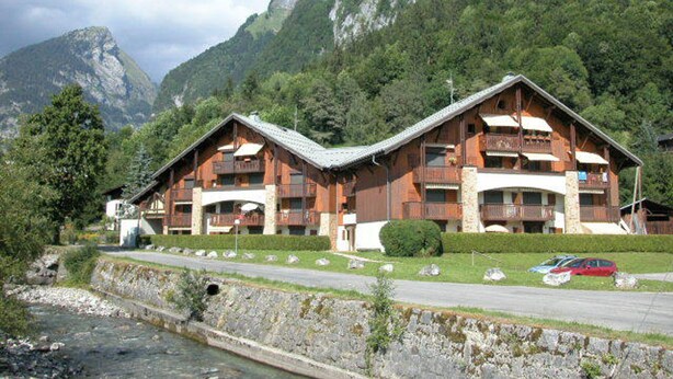 Un rifugio di montagna con numerosi balconi e facciata in pietra, situato vicino a un fiume e circondato da montagne boscose.