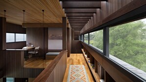 Un interior moderno con un largo pasillo, paredes de madera, un escritorio con una silla y un amplio ventanal con vistas a árboles.