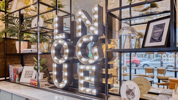 A reception desk with illuminated letters spelling 'Lounge,' a framed award, and a view of the street outside. - Hub By Premier Inn London Clerkenwell (London)