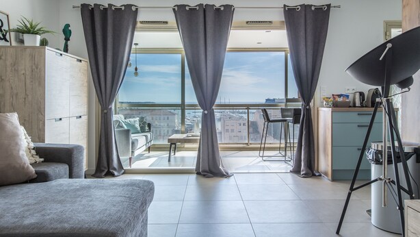 A modern living room with a sofa, a dining area, and a balcony with a city view. - Admire Panoramic Ocean Views Thru Walls of Windows (Cannes)