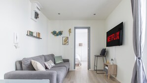 Ein modernes Wohnzimmer mit einem grauen Sofa, einem an der Wand befestigten Fernseher, der 'NETFLIX' anzeigt, einem kleinen Holztisch und einer Tür, die in einen anderen Raum führt.