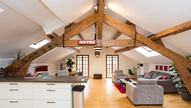 Un loft spacieux avec de hauts plafonds, des poutres apparentes, un coin cuisine, un espace repas et un salon doté de canapés et d’une table basse.