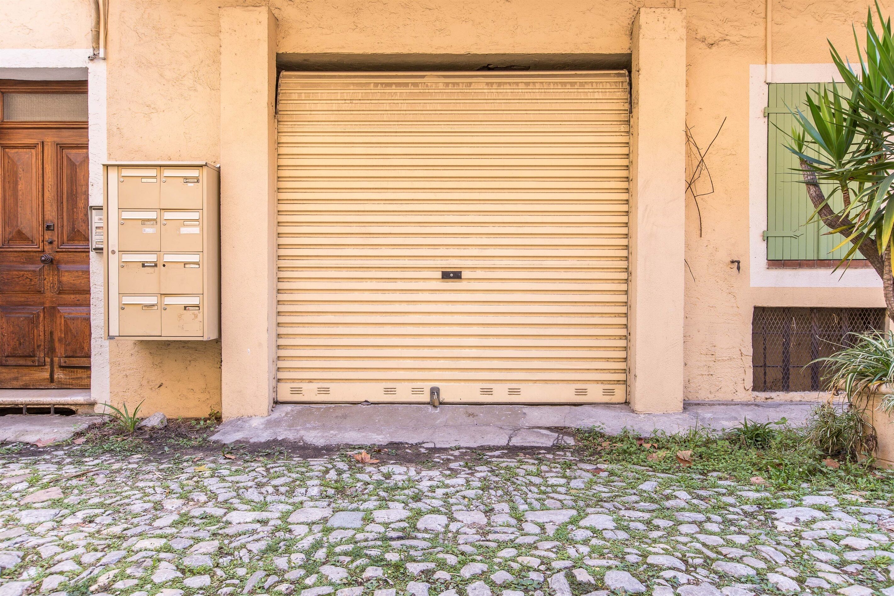Un edificio con un portone beige chiuso dotato di persiana a rullo, una porta in legno sulla sinistra e una cassetta postale sullo stesso lato.