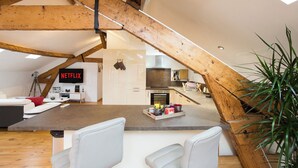 Une cuisine moderne avec des poutres en bois, un coin repas avec des chaises blanches et un écran de télévision bien visible affichant le logo « NETFLIX ».