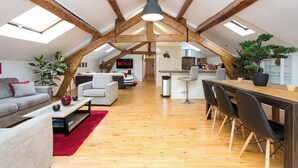 Un loft spacieux avec un plafond haut, des poutres en bois, un coin repas avec une longue table et des chaises, un salon avec un canapé et une table basse, et une cuisine à l’arrière-plan.