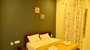 A hotel room with a bed, a bedside table with a phone and a glass, and a window with floral curtains. - Nilton Bay Residency (Tindivanam)
