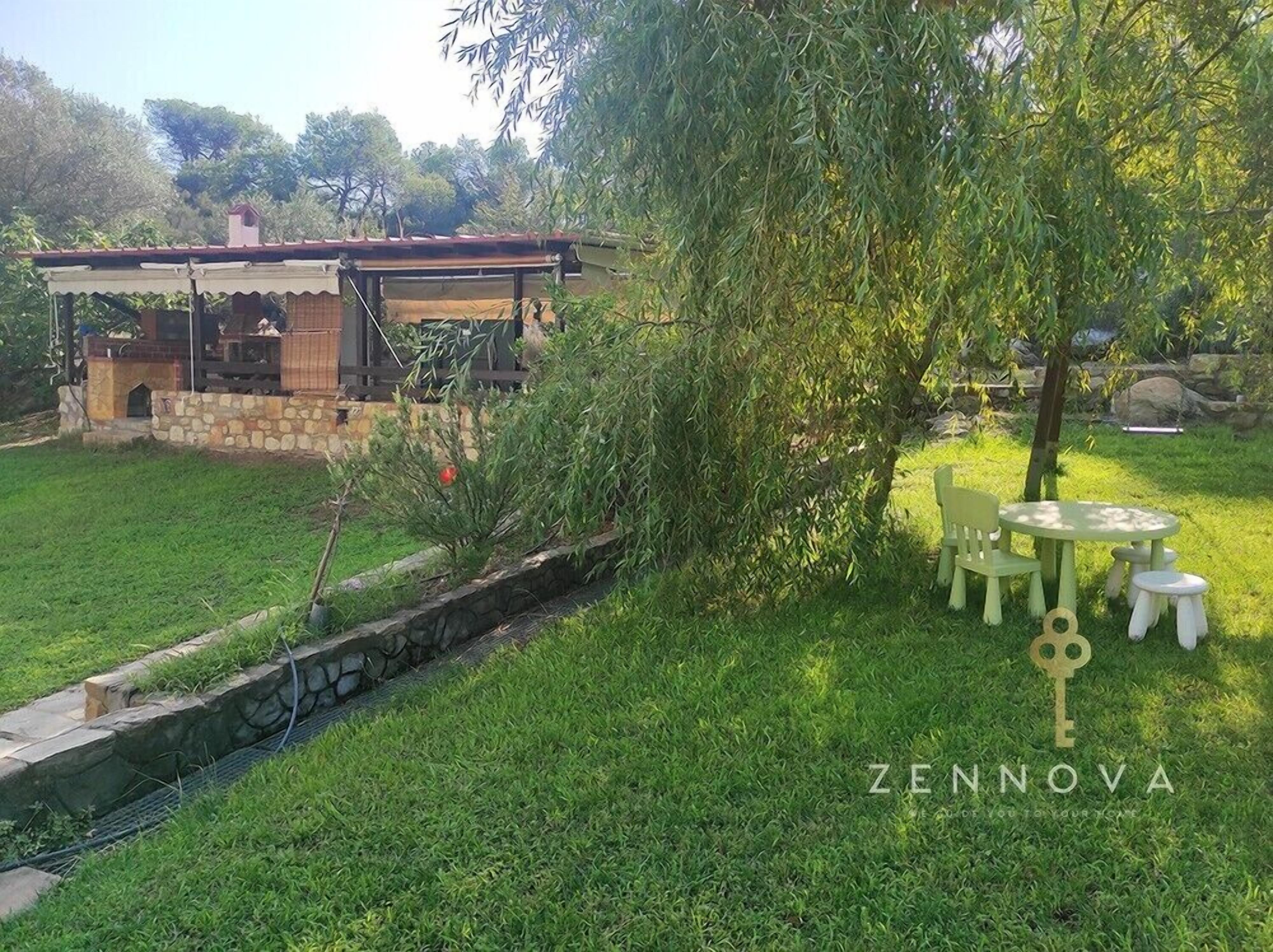 Une maison avec des fondations en pierre, un patio couvert et un petit ruisseau. On y trouve un arbre aux feuilles vertes et une table avec des chaises dans la cour.