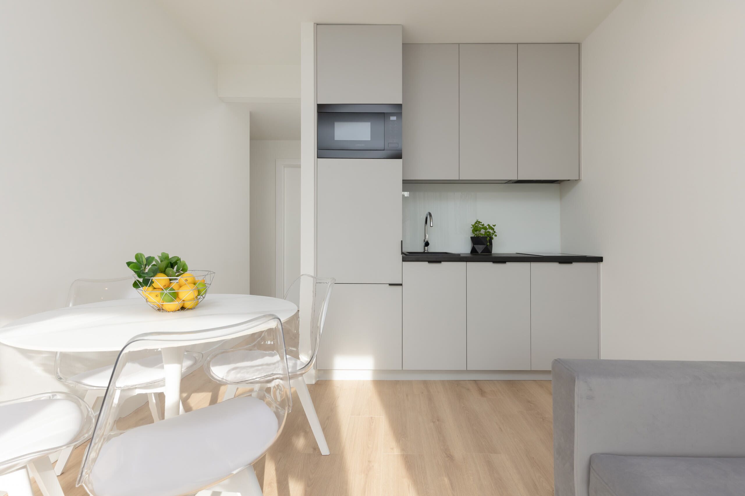 A compact kitchen with white cabinets, a microwave, and a small countertop. There is a dining table with chairs and a bowl of fruit.