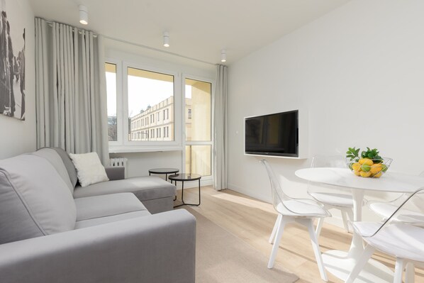 A modern living room with a grey sofa, a flat-screen TV, a dining table with chairs, and a window with curtains.