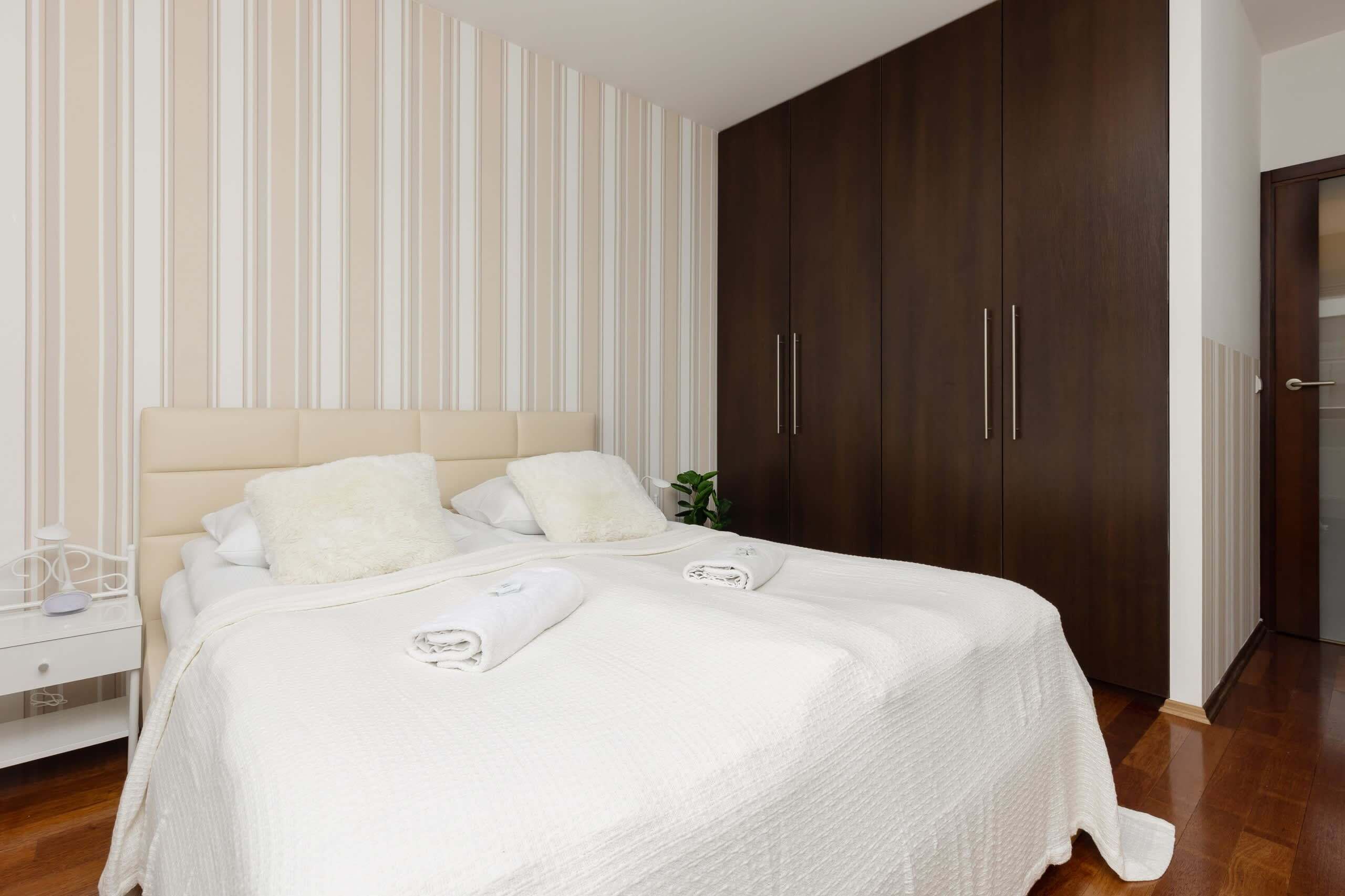 A neatly made bed with white linens and pillows, a bedside table with a lamp, and a dark wooden wardrobe with silver handles.