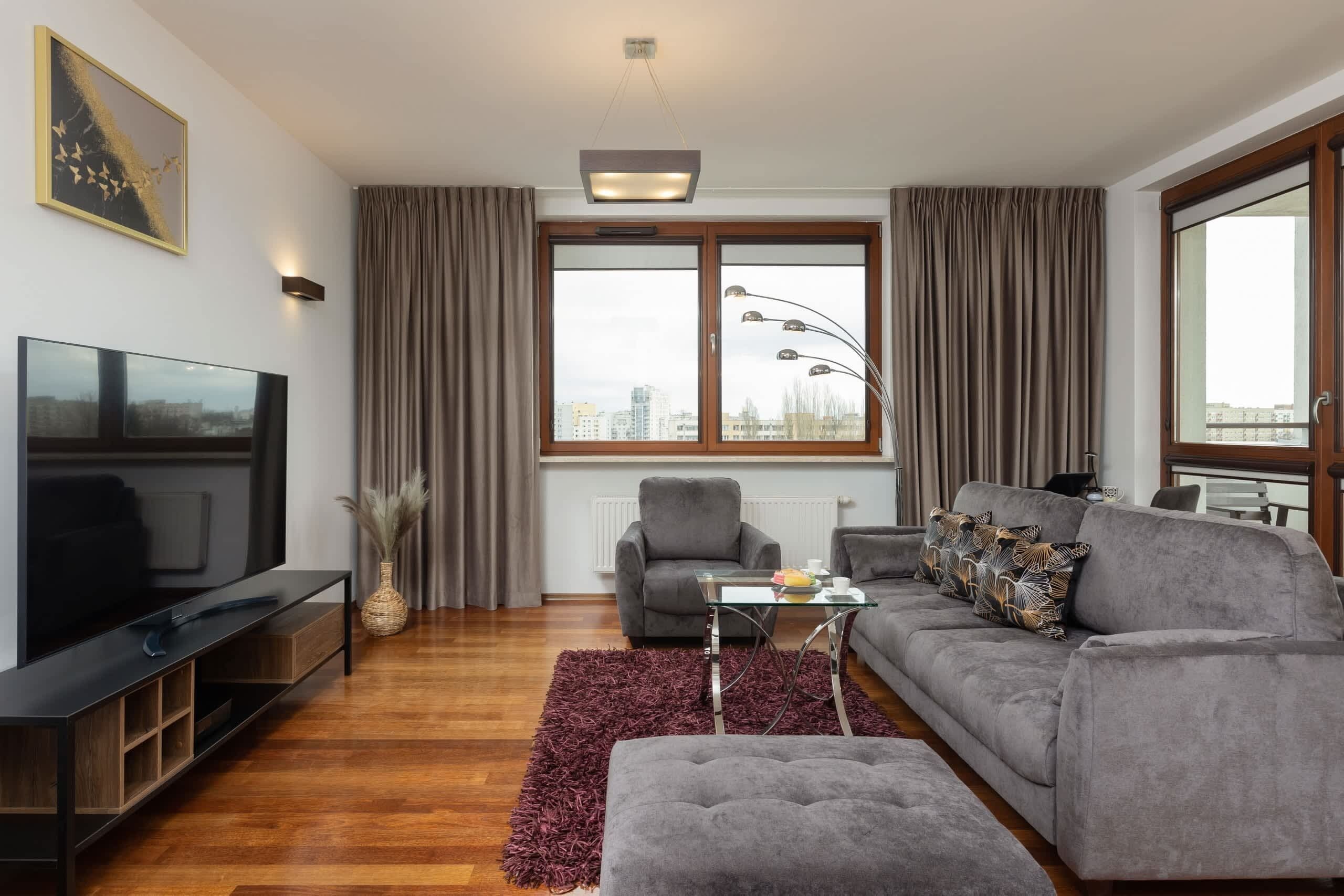 A modern living room with a flat-screen TV, grey sofa, and a large window with curtains.
