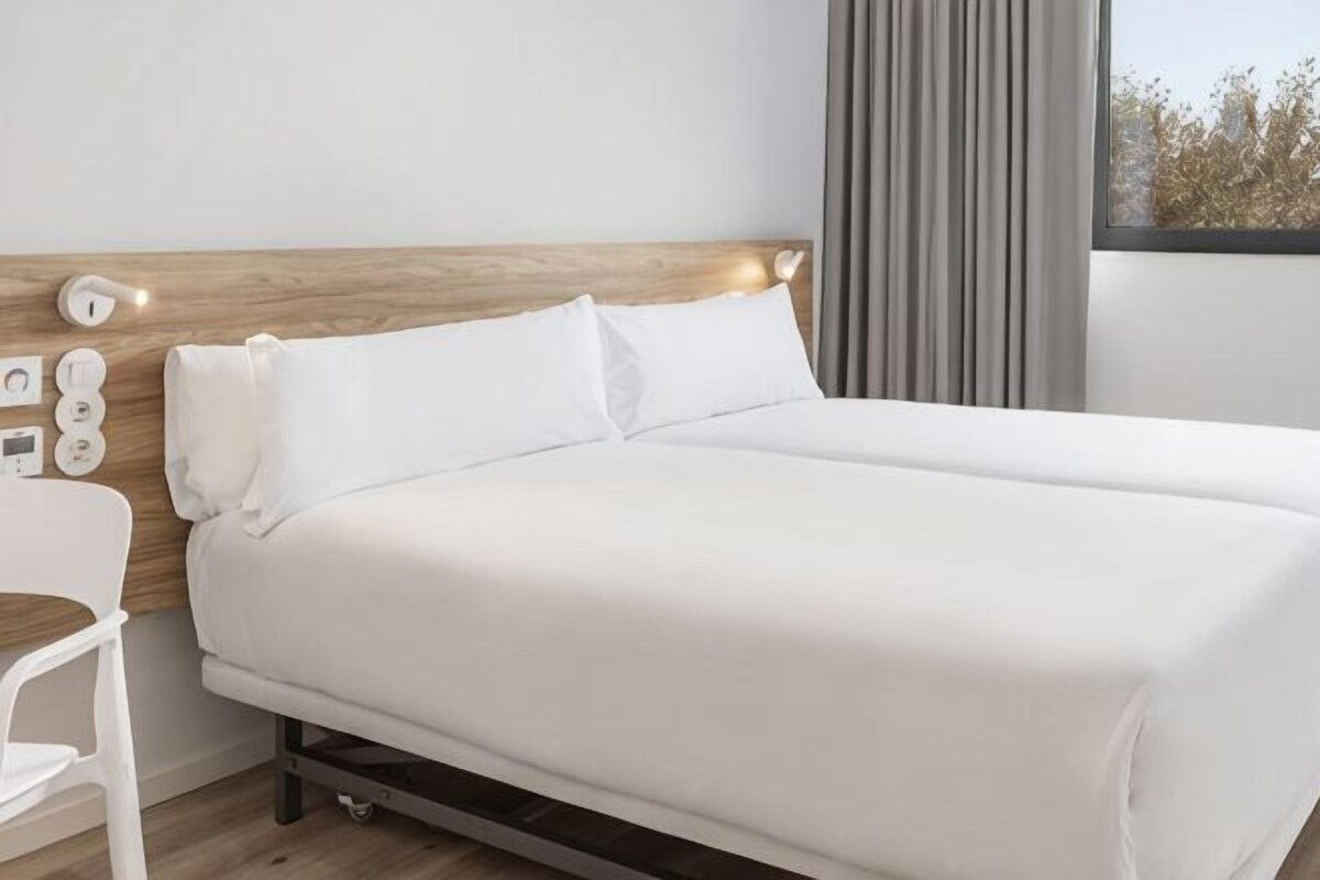 A single bed with a wooden headboard, white bedding, a bedside table, and a window with curtains.