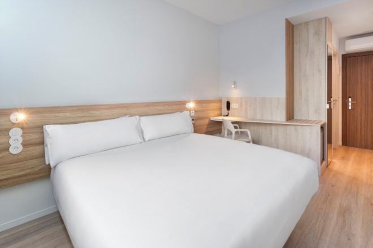 A modern hotel room with a large bed, a wooden headboard, a small desk, and a wardrobe.