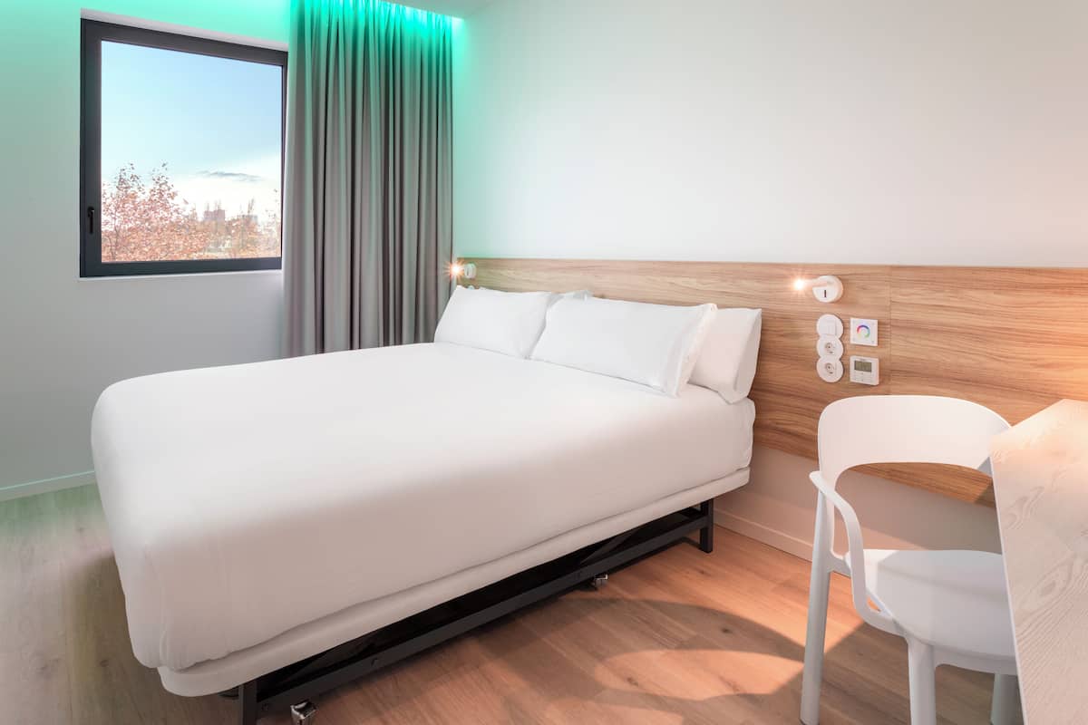 Une chambre d’hôtel moderne avec un grand lit, une tête de lit en bois, une fenêtre avec des rideaux et une chaise blanche.