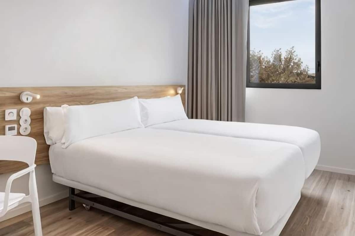 Un lit bien fait dans une chambre dotée d’une tête de lit en bois, d’une fenêtre avec des rideaux et d’une chaise blanche.