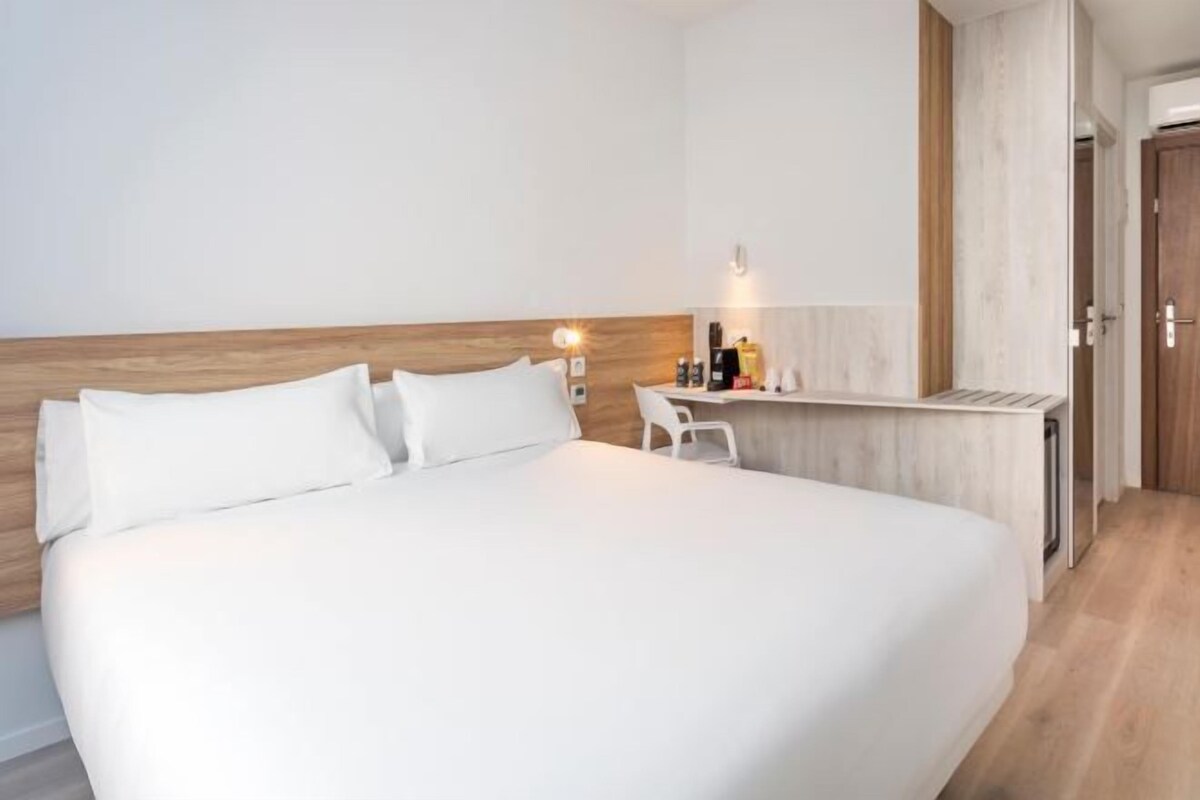 A modern hotel room with a large bed, a small desk, and a wooden headboard.