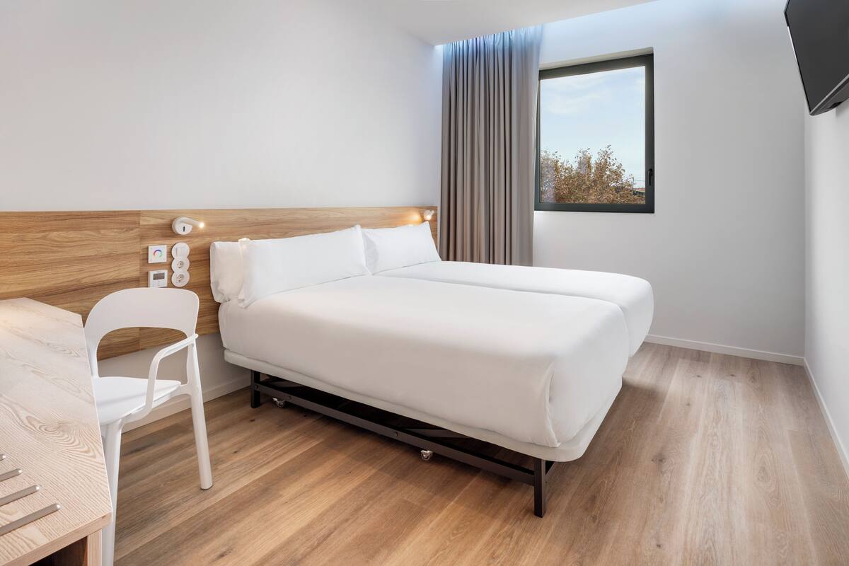 Une chambre d’hôtel moderne avec une tête de lit en bois, un lit simple, une chaise blanche et une fenêtre avec des rideaux.