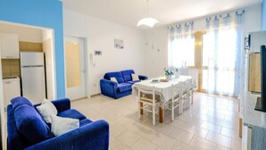 A living room with a blue sofa, a dining table with chairs, and a kitchen area with a refrigerator. - CASAMARiNATSG- Sea View (Torre San Giovanni)