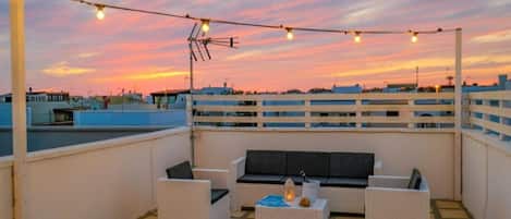 Terraza en la azotea con mobiliario exterior, luces colgantes y vistas del horizonte urbano al atardecer.