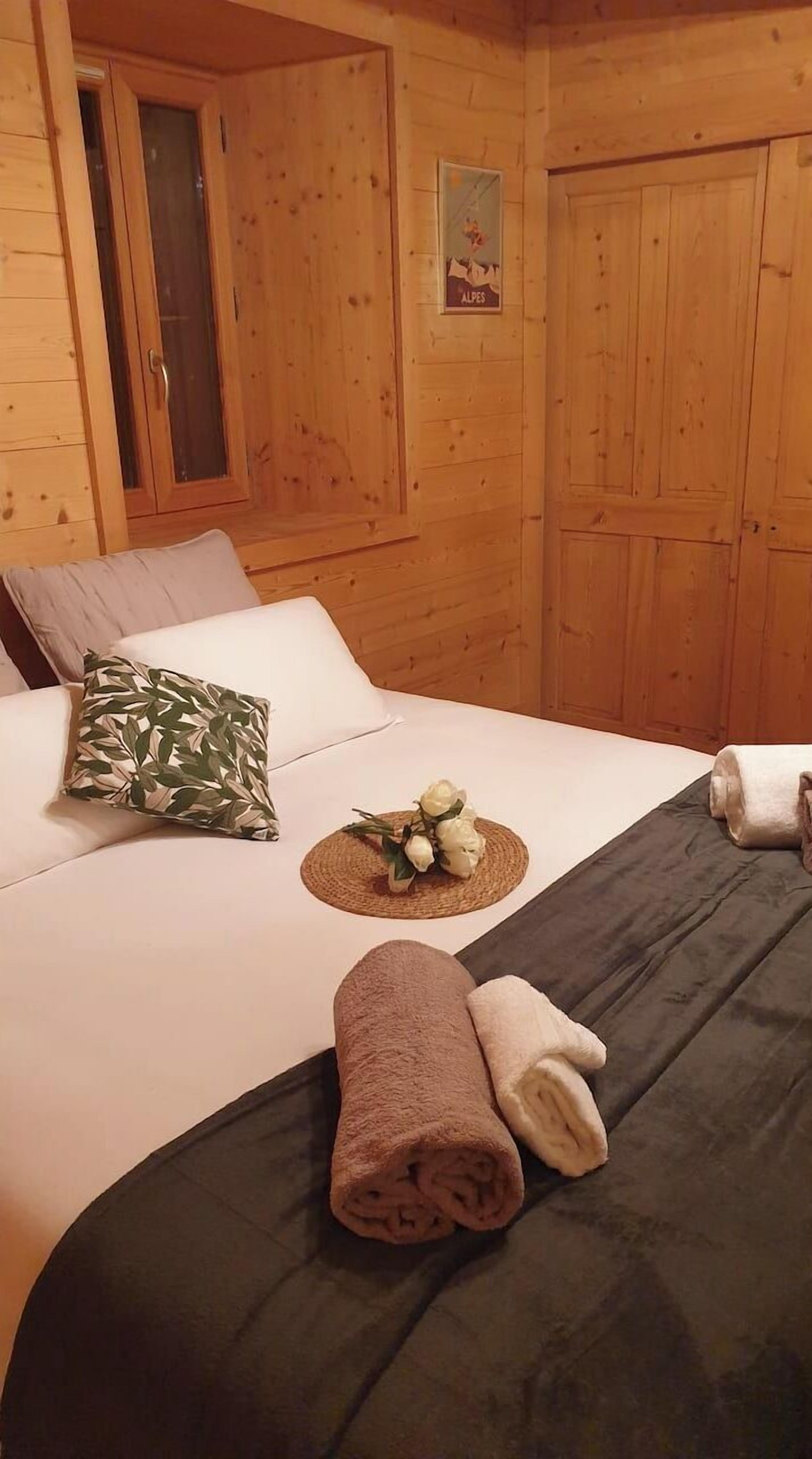 Habitación de cabaña de madera con una cama, almohadas, una toalla enrollada y una mesita con un arreglo floral.