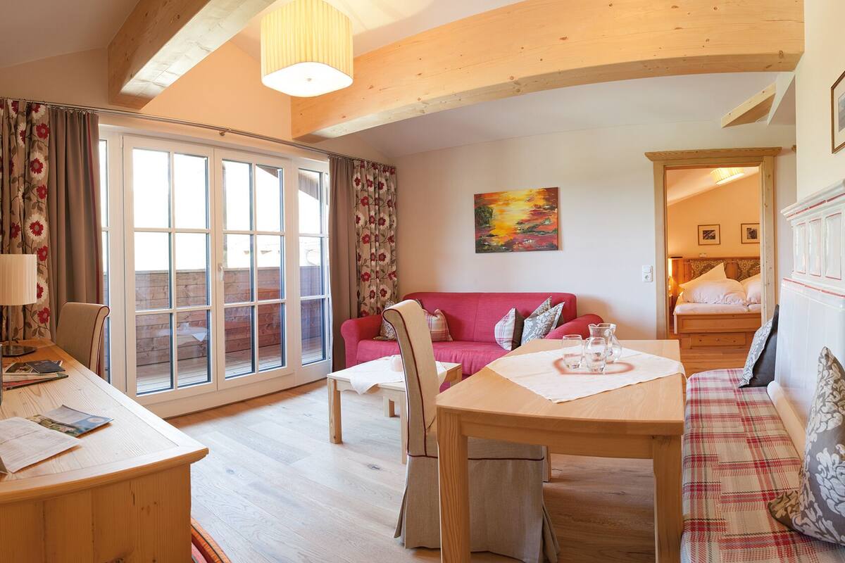 A cozy living room with a wooden dining table, a red sofa, and a large window with curtains.