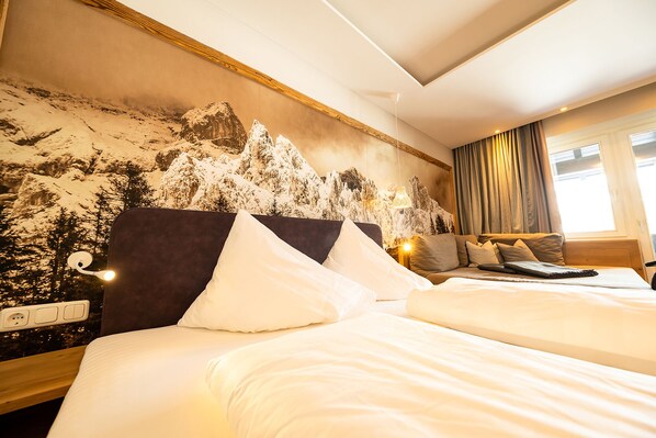 A hotel room with two beds, a mountain mural on the wall, a sofa, and a window with curtains.