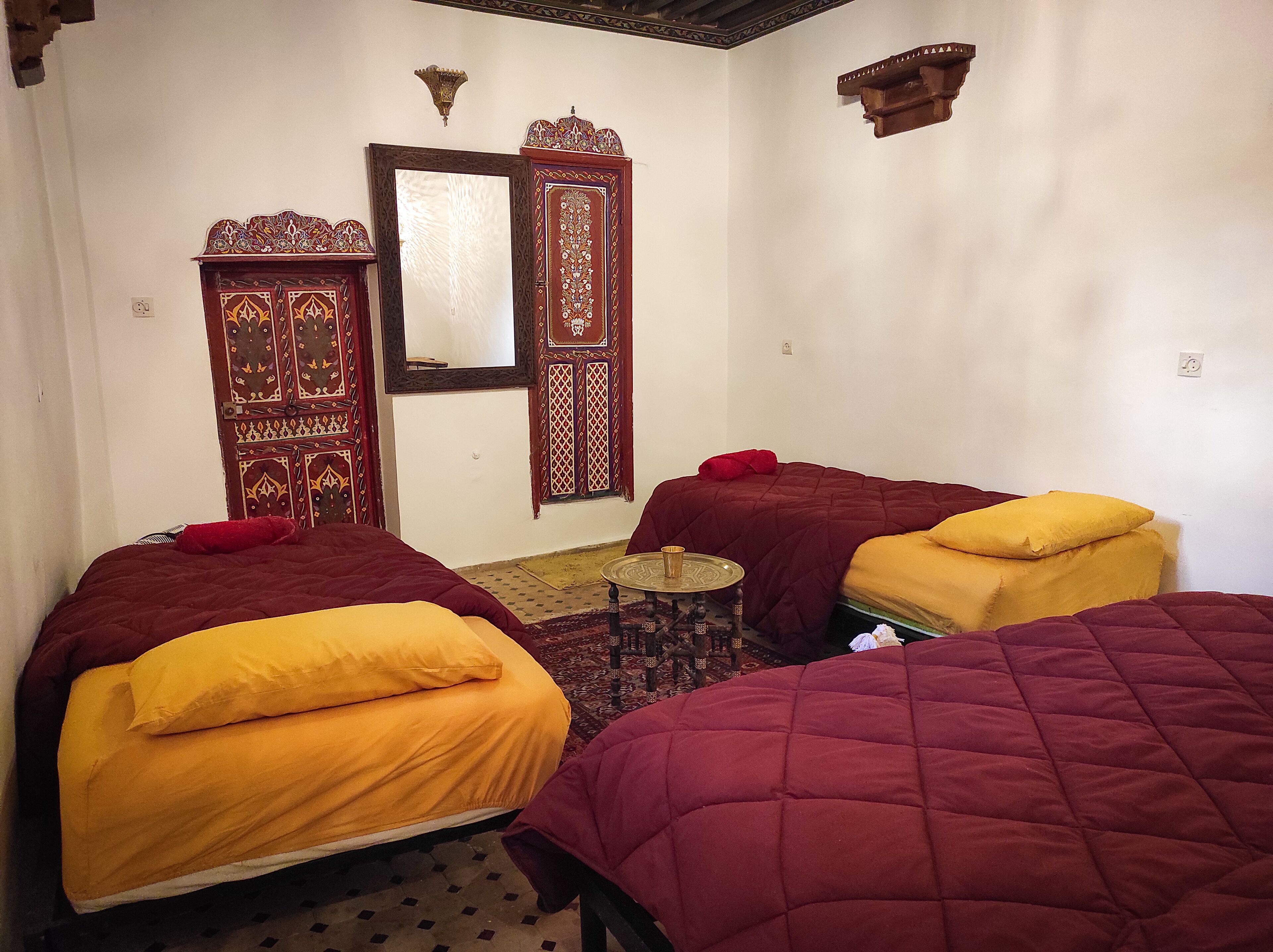 Una habitación con dos camas, cada una con una colcha roja y amarilla, una pequeña mesa redonda con una bandeja y un espejo decorativo en la pared.