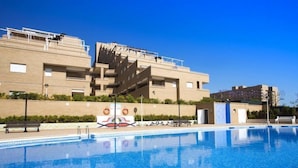 A swimming pool with a clear blue water surface, surrounded by a tiled deck. In the background, there is a multi-story building with a modern architectural design, featuring balconies and a flat roof. - Jardines del Mar I by Interhome (Oropesa del Mar)