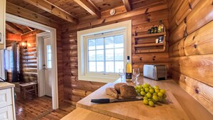 A wooden cabin kitchen with a large window, a wooden countertop, a toaster, a bottle of wine, and a bowl of grapes.