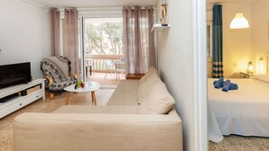 A cozy living area with a sofa, a TV, and a small table with a fruit bowl. There is a balcony with a view of trees. - cye 6 Cap Salou by Interhome (Salou)