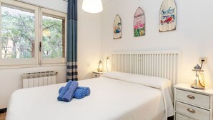 A neatly made bed with rolled towels, a white radiator, a window with curtains, and wall decorations including surfboards. - cye 6 Cap Salou by Interhome (Salou)