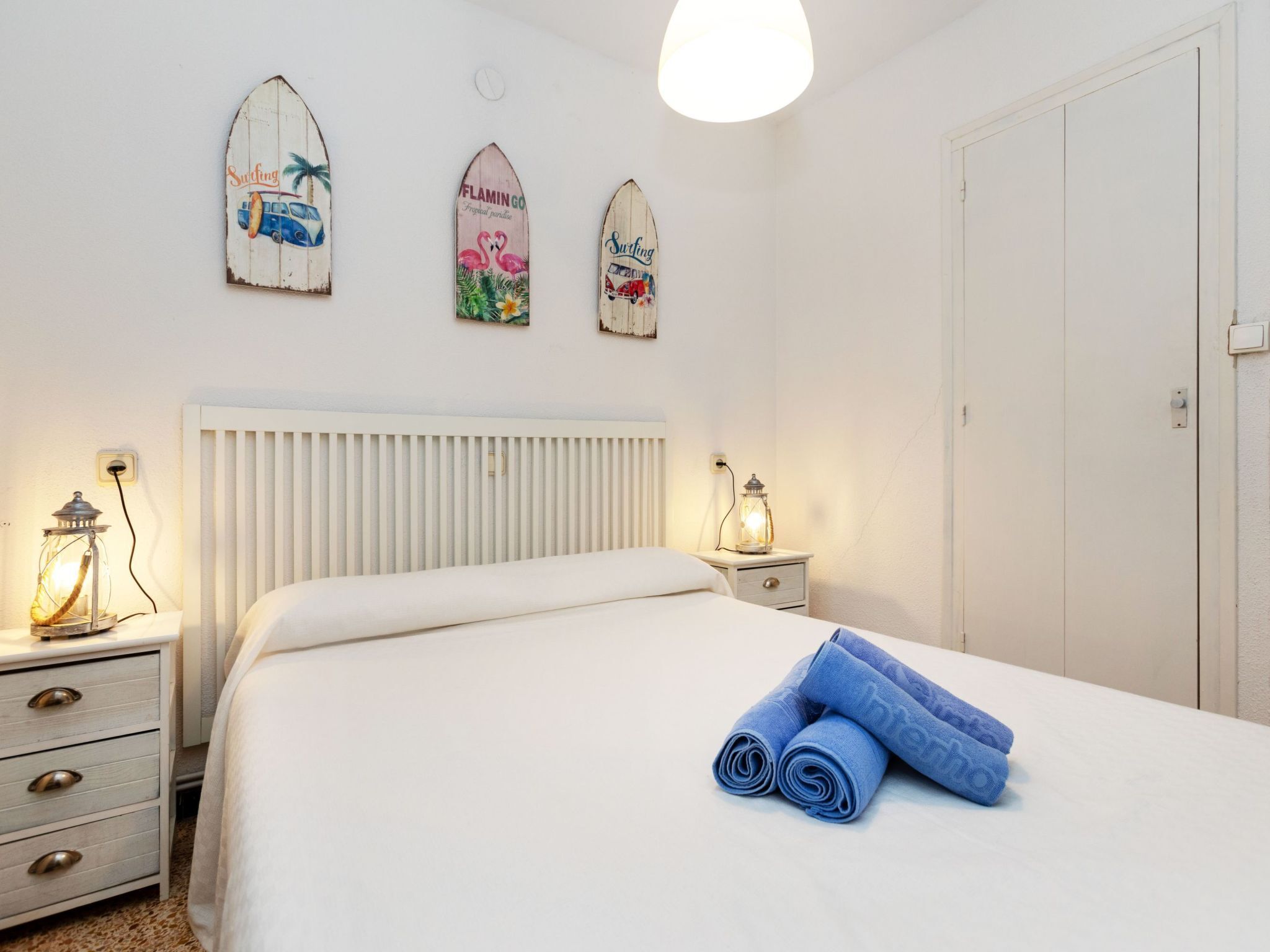 Een slaapkamer met een bed, een nachtkastje met een lamp, en drie surfplanken aan de muur.