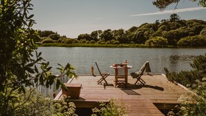 En træterrasse med et bord og stole, med udsigt over en sø, omgivet af grøn bevoksning.