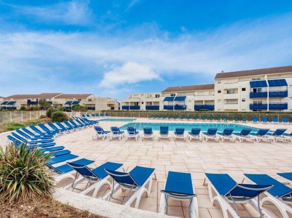 A pool area with lounge chairs arranged in a semi-circle around a central pool, surrounded by multi-story buildings with balconies. - Bleu Marine by Interhome (Lacanau)
