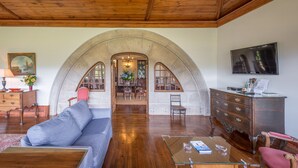 Un salón amplio con techo de madera, un arco de piedra que conduce a un comedor y un televisor montado en la pared.