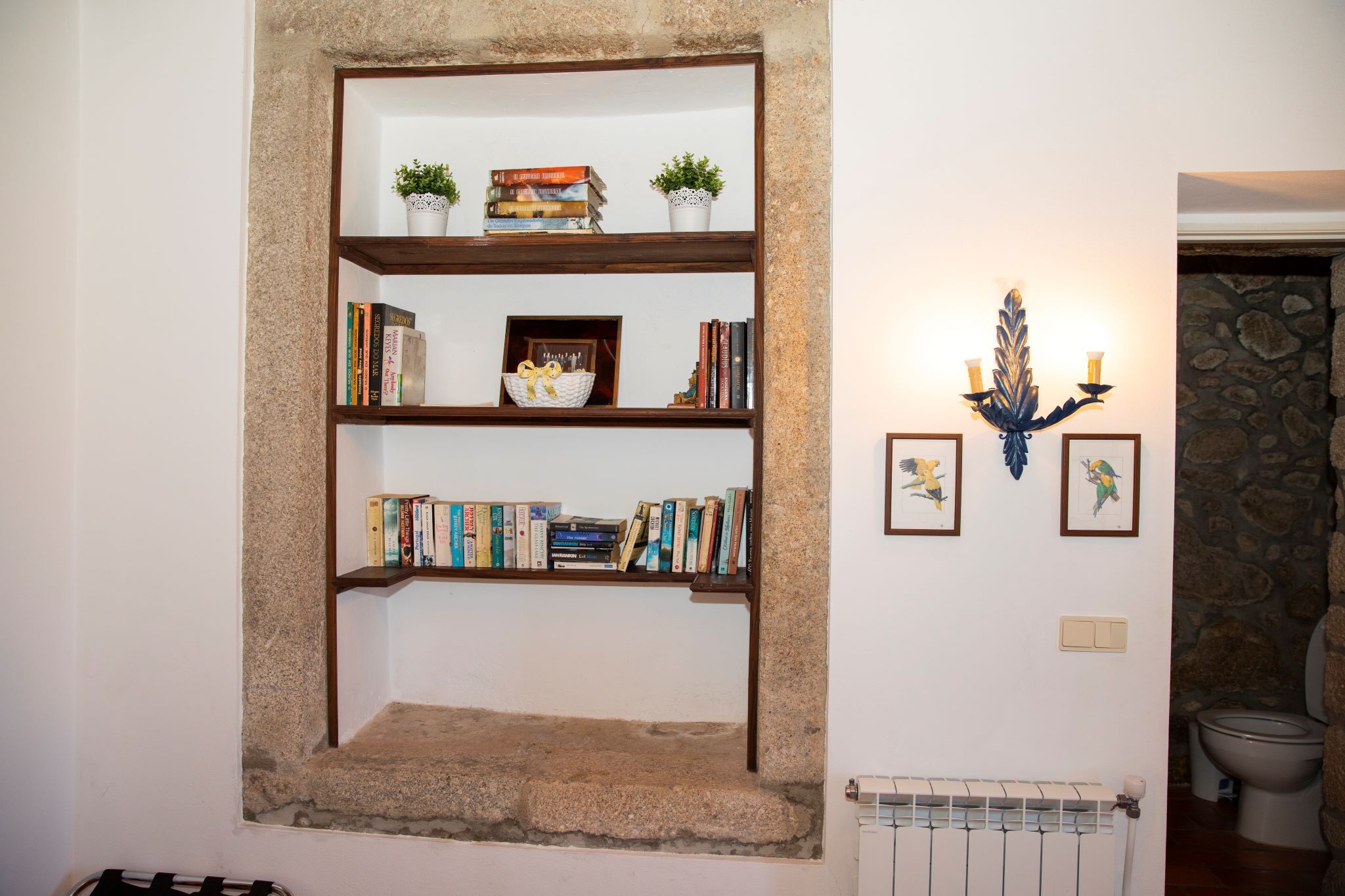 A bookshelf with books and decorative items, a wall-mounted light fixture, and a stone wall in the background.