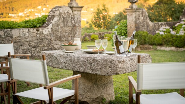 A stone table set with plates, glasses, and a bottle, surrounded by chairs in a garden with stone walls and a view of a village in the background. - Casa do Tanque at Quinta de Luou (Ponte de Lima)