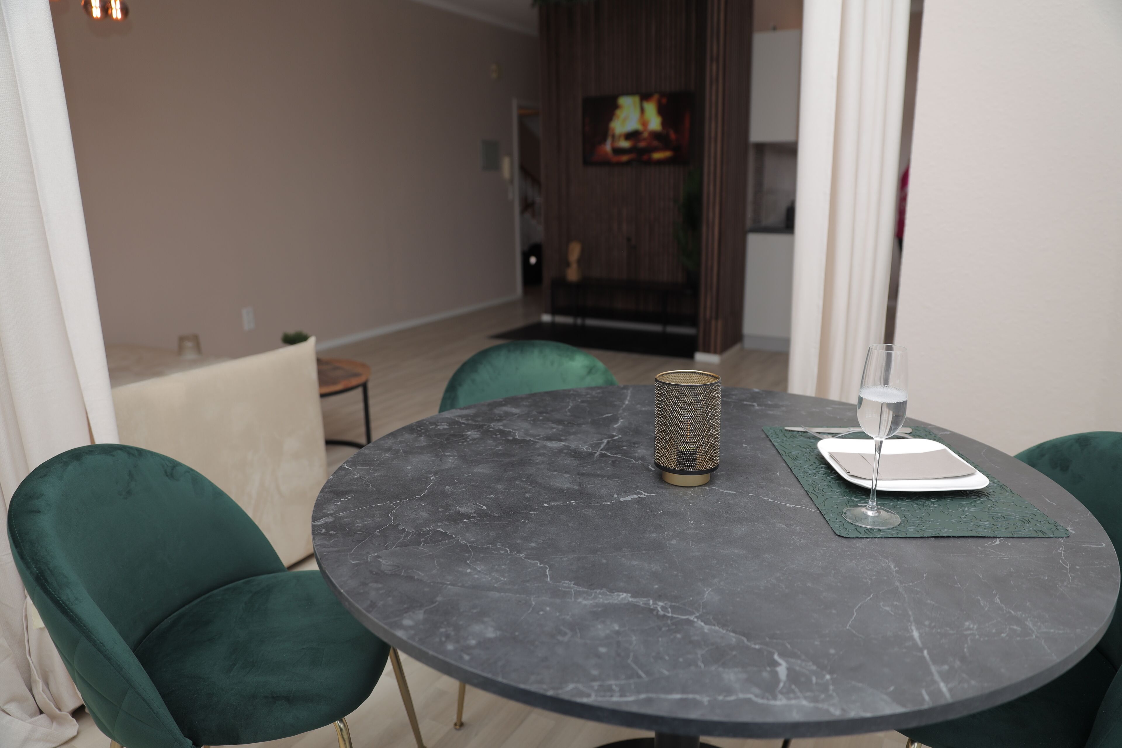 Una mesa redonda con cubierta de mármol, rodeada por dos sillas verdes. Una pequeña lámpara de mesa y una copa de vino están sobre la mesa.