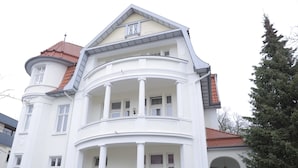 A white multi-story building with a curved balcony, a tower with a red roof, and a large evergreen tree in front.
