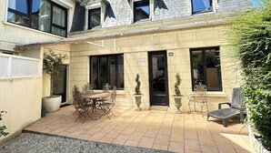 A two-story house with a tiled patio, a dining table with chairs, and a small garden area.