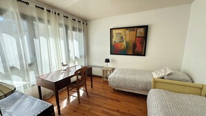 A room with a wooden desk, a chair, a bed with patterned bedding, a small table with a lamp, and a painting on the wall.