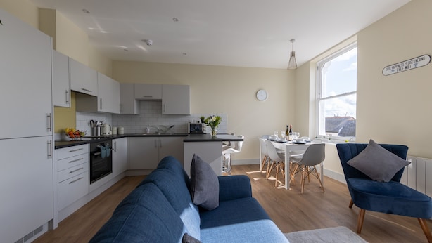 Una cocina moderna con armarios blancos, una zona de comedor con mesa y sillas, y un sofá azul en una sala de estar.