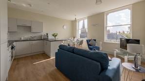 Una cocina moderna con armarios blancos, un comedor con mesa y sillas, un sofá azul y un amplio ventanal con vistas a edificios.