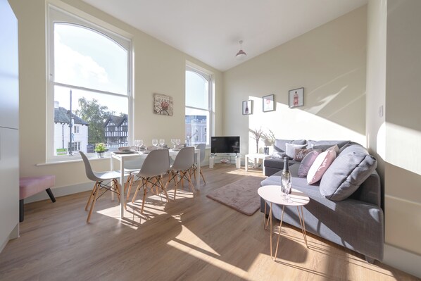 A modern living room with a dining area, large windows, a flat-screen TV, and a grey sofa.
