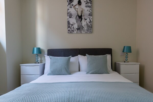 A neatly made bed with a light blue blanket and pillows, two bedside tables with teal lamps, and a wall-mounted painting above the bed.
