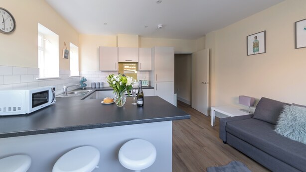 A modern kitchen with a central island, white bar stools, a grey sofa, and a small white table.